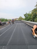 Three people are working on a flat rooftop, applying a black material to the surface. One person is kneeling and spreading the material with a tool; another is walking away while carrying something, and the third person is seated to the side. Various tools and supplies are scattered around, and trees are visible in the background.
