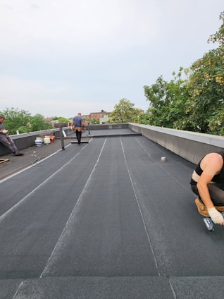 Three people are working on a flat rooftop, applying a black material to the surface. One person is kneeling and spreading the material with a tool; another is walking away while carrying something, and the third person is seated to the side. Various tools and supplies are scattered around, and trees are visible in the background.