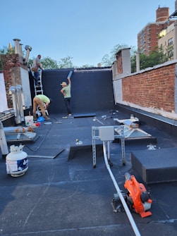 A rooftop workspace where three individuals are engaged in construction or renovation activities. One person is on a ladder working on a wall or chimney, another is standing and holding a black panel, and a third person is bent over handling tools or equipment. Nearby are various construction materials, tools like a leaf blower, a propane tank, and some ductwork. The surrounding environment includes brick walls and a view of trees and buildings in the background.