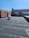 A flat rooftop area with a dark surface and a few objects, including a vent pipe and a small utility structure. The surrounding structures are made of red brick and feature large windows. There is a clear blue sky above, and a slight shadow is cast across the rooftop.