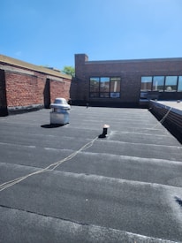 A flat rooftop area with a dark surface and a few objects, including a vent pipe and a small utility structure. The surrounding structures are made of red brick and feature large windows. There is a clear blue sky above, and a slight shadow is cast across the rooftop.