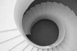 An aerial view of a spiral staircase with a smooth, continuous curve forming the steps and railing. The staircase is predominantly white, creating a contrasting shadow effect in the center.