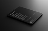 A sleek, black tablet-like object rests on a dark surface, displaying illuminated text in a minimalist style. The text includes features and information about a mock-up, primarily in white font, conveying a modern and professional look.