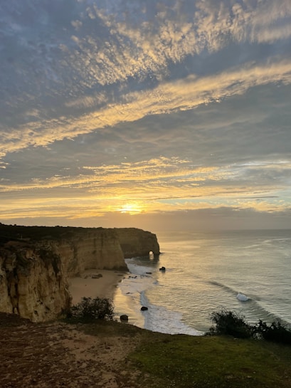 A scenic view of the ocean with a golden sunset on the horizon. Rocky cliffs extend into the water with a natural arch formation visible. The sky is filled with wispy clouds reflecting the warm hues of the setting sun. Vegetation is sparse and situated atop the rugged cliffside overlooking the beach and gently crashing waves.
