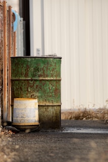 A weathered, green rusted metal barrel stands on a pavement next to a yellow and blue bucket. The background features a corrugated metal wall, and the scene is framed by a rusty chain-link fence on the left.