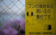 A yellow sign features cartoon illustrations of a dog holding a bag, along with Japanese text indicating the responsibility of pet owners. The sign is seen against a backdrop of chain-link fencing and blurred purple flowers.