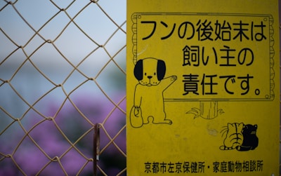 A yellow sign features cartoon illustrations of a dog holding a bag, along with Japanese text indicating the responsibility of pet owners. The sign is seen against a backdrop of chain-link fencing and blurred purple flowers.