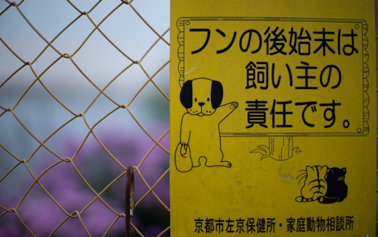 A yellow sign features cartoon illustrations of a dog holding a bag, along with Japanese text indicating the responsibility of pet owners. The sign is seen against a backdrop of chain-link fencing and blurred purple flowers.