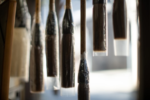 A series of large paintbrushes hang vertically, enclosed in plastic wrapping. The focus is on the bristles, with a soft, out-of-focus background providing contrast.