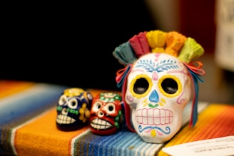 Three decorative skulls, known as sugar skulls, rest on a multicolored blanket. The largest skull is adorned with intricate floral patterns in vibrant colors and has colorful paper flowers on top. The two smaller skulls, one blue and the other red, follow a similar style with painted designs and patterns.