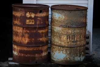 Two rusty metal barrels stand side by side on a concrete surface. One barrel has a predominantly dark brown rust while the other displays a mix of rust and faded blue paint. Both barrels show signs of weathering and corrosion, with old and illegible text or symbols barely visible through the rust.