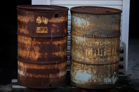 Two rusty metal barrels stand side by side on a concrete surface. One barrel has a predominantly dark brown rust while the other displays a mix of rust and faded blue paint. Both barrels show signs of weathering and corrosion, with old and illegible text or symbols barely visible through the rust.