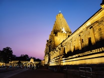 A beautifully lit ancient temple with intricate carvings, set against a dramatic evening sky with hues of purple and blue. There are people walking around the temple grounds, adding a sense of scale and liveliness.