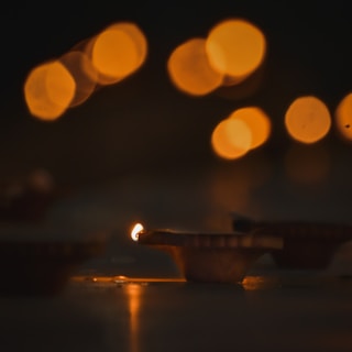 A warm glowing diya lamp is in focus, casting a gentle light. The background features soft, circular bokeh effects with a warm, golden hue, creating a serene ambiance.