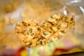 A close-up view inside an opened bag of crispy snacks, likely chips or a similar savory treat. The chips are seasoned with green herbs and spices, with a mix of yellow and brown hues indicating the flavoring. The bag's inner surface can be partially seen, reflecting light and creating a shiny effect.
