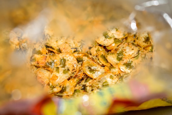 A close-up view inside an opened bag of crispy snacks, likely chips or a similar savory treat. The chips are seasoned with green herbs and spices, with a mix of yellow and brown hues indicating the flavoring. The bag's inner surface can be partially seen, reflecting light and creating a shiny effect.