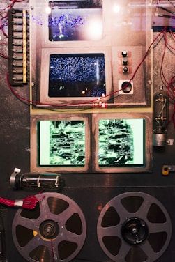 A vintage piece of machinery features multiple screens displaying abstract patterns, surrounded by various buttons, switches, and wires. Two large film reels are positioned at the bottom, adding to the retro technological aesthetic. The overall setup suggests an analog device possibly from the mid-20th century.