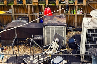A cluttered outdoor space with a skeleton sitting on a patio chair. Surrounding the skeleton are various neglected objects including rusty air conditioning units, gardening tools hanging on a wooden fence, and scattered leaves on the ground.