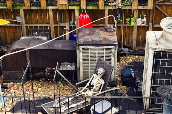 A cluttered outdoor space with a skeleton sitting on a patio chair. Surrounding the skeleton are various neglected objects including rusty air conditioning units, gardening tools hanging on a wooden fence, and scattered leaves on the ground.