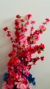 A vibrant arrangement of artificial flowers with delicate pink and red blossoms, interspersed with green leaves, against a neutral background. The flowers are intricately detailed, capturing various shades, with some blue blossoms at the base adding contrast.