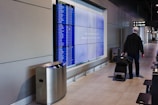A person is walking through an airport terminal with a rolling suitcase. The individual is passing in front of a flight information display showing numerous departure details. The terminal has a modern design, featuring a line of chairs and overhead signage indicating gate directions.
