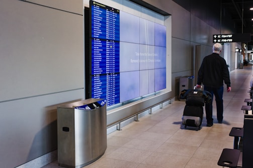 A person is walking through an airport terminal with a rolling suitcase. The individual is passing in front of a flight information display showing numerous departure details. The terminal has a modern design, featuring a line of chairs and overhead signage indicating gate directions.