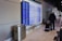 A person is walking through an airport terminal with a rolling suitcase. The individual is passing in front of a flight information display showing numerous departure details. The terminal has a modern design, featuring a line of chairs and overhead signage indicating gate directions.