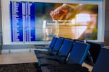 A modern airport seating area with several blue seats and two large screens. One screen displays a flight information board with departure details in a grid. The other screen has an advertisement showcasing a drink being poured into a glass with text in both English and French. The area is well-lit and appears clean and organized.