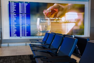 A modern airport seating area with several blue seats and two large screens. One screen displays a flight information board with departure details in a grid. The other screen has an advertisement showcasing a drink being poured into a glass with text in both English and French. The area is well-lit and appears clean and organized.