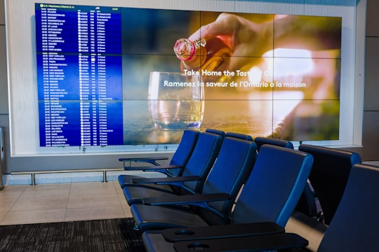 A modern airport seating area with several blue seats and two large screens. One screen displays a flight information board with departure details in a grid. The other screen has an advertisement showcasing a drink being poured into a glass with text in both English and French. The area is well-lit and appears clean and organized.