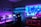 A vibrant gaming setup featuring multiple monitors displaying a cyberpunk cityscape. The room is illuminated with neon purple and blue LED lights, adding a futuristic ambiance. A custom-built PC with glowing components sits to the side, and shelves with collectible figurines are visible. A large digital screen on the wall shows an animated city scene, enhancing the immersive atmosphere.