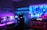 A vibrant gaming setup featuring multiple monitors displaying a cyberpunk cityscape. The room is illuminated with neon purple and blue LED lights, adding a futuristic ambiance. A custom-built PC with glowing components sits to the side, and shelves with collectible figurines are visible. A large digital screen on the wall shows an animated city scene, enhancing the immersive atmosphere.