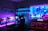 A vibrant gaming setup featuring multiple monitors displaying a cyberpunk cityscape. The room is illuminated with neon purple and blue LED lights, adding a futuristic ambiance. A custom-built PC with glowing components sits to the side, and shelves with collectible figurines are visible. A large digital screen on the wall shows an animated city scene, enhancing the immersive atmosphere.