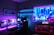 A vibrant gaming setup featuring multiple monitors displaying a cyberpunk cityscape. The room is illuminated with neon purple and blue LED lights, adding a futuristic ambiance. A custom-built PC with glowing components sits to the side, and shelves with collectible figurines are visible. A large digital screen on the wall shows an animated city scene, enhancing the immersive atmosphere.