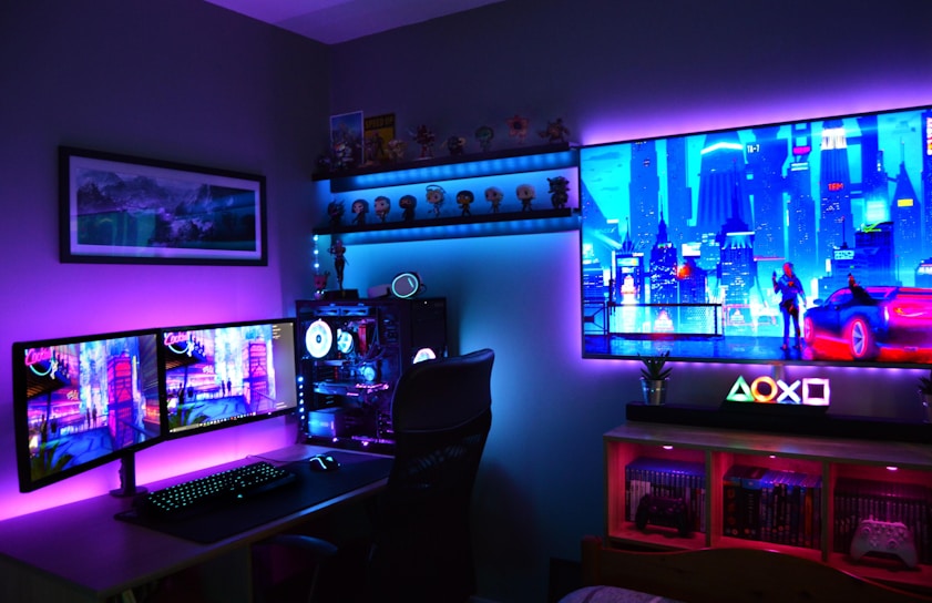 A vibrant gaming setup featuring multiple monitors displaying a cyberpunk cityscape. The room is illuminated with neon purple and blue LED lights, adding a futuristic ambiance. A custom-built PC with glowing components sits to the side, and shelves with collectible figurines are visible. A large digital screen on the wall shows an animated city scene, enhancing the immersive atmosphere.