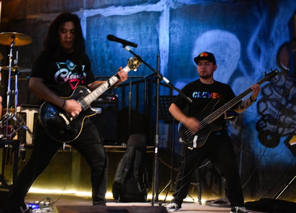 Two musicians are performing on a stage with a vibrant, graffiti-covered backdrop. One is playing an electric guitar, while the other is on a bass guitar. They are both dressed casually, and the setting suggests an energetic and lively atmosphere typical of a live music venue. Musical equipment like amps and a microphone stand are visible.