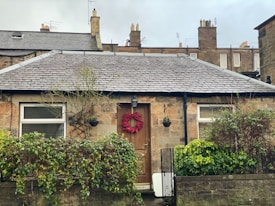 A quaint stone cottage with a dark gray roof, featuring a wooden door adorned with a red wreath. Framed windows flanking the door display drawn blinds. In front of the house, a lush green hedge and several plants fill the small garden, with a stone wall and black railing at the front.