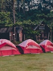 Red and white tents are set up on a grassy area near a wooded backdrop. The tents are arranged in a line, in front of rustic wooden cabins. String lights are hung between trees, creating a cozy atmosphere among the lush green foliage.