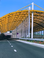 An empty road leading into a tunnel with a modern design. The tunnel entrance is adorned with a yellow geometric structure, and there are traffic signs and green foliage lining the roadway.