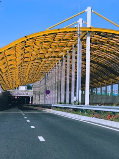 An empty road leading into a tunnel with a modern design. The tunnel entrance is adorned with a yellow geometric structure, and there are traffic signs and green foliage lining the roadway.