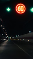 A dimly lit tunnel with a red circular speed limit sign displaying 60 near the ceiling. Green arrows point downward, and the tunnel has evenly spaced environmental lights along the walls.
