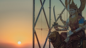 A statue of a goddess adorned with traditional attire and jewelry is positioned behind a pane of glass, bathed in the warm glow of a sunset. The figure holds a sword and other symbolic items, and sits atop a stylized lion. The background features the sun setting on the horizon, casting a serene and mystical atmosphere.