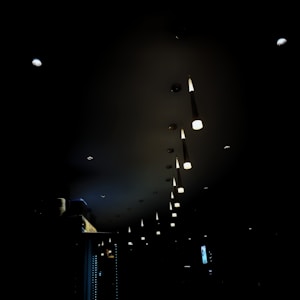 A dimly lit interior featuring a row of pendant lights hanging from the ceiling. The lights are in a linear arrangement, casting a warm glow in an otherwise dark environment. The setting appears to be a modern space, possibly a restaurant or a bar, with minimal visible furnishing and fixtures.