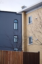 Two residential buildings are shown side by side, one with a grey façade featuring arched and rectangular windows, and the other with yellowish brickwork and rectangular windows. A bare tree with branches is in the foreground, along with a wooden fence.