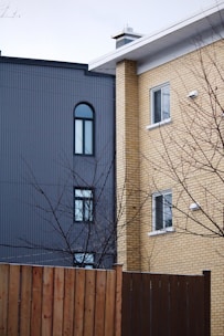Two residential buildings are shown side by side, one with a grey façade featuring arched and rectangular windows, and the other with yellowish brickwork and rectangular windows. A bare tree with branches is in the foreground, along with a wooden fence.