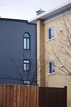 Two residential buildings are shown side by side, one with a grey façade featuring arched and rectangular windows, and the other with yellowish brickwork and rectangular windows. A bare tree with branches is in the foreground, along with a wooden fence.
