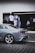 A sleek grey car is parked on a cobblestone street in front of a clothing shop displaying white embroidered shirts and dresses. The foreground features an intricate pavement pattern, and the background shows the entrance to a store with a dark interior and hanging garments.