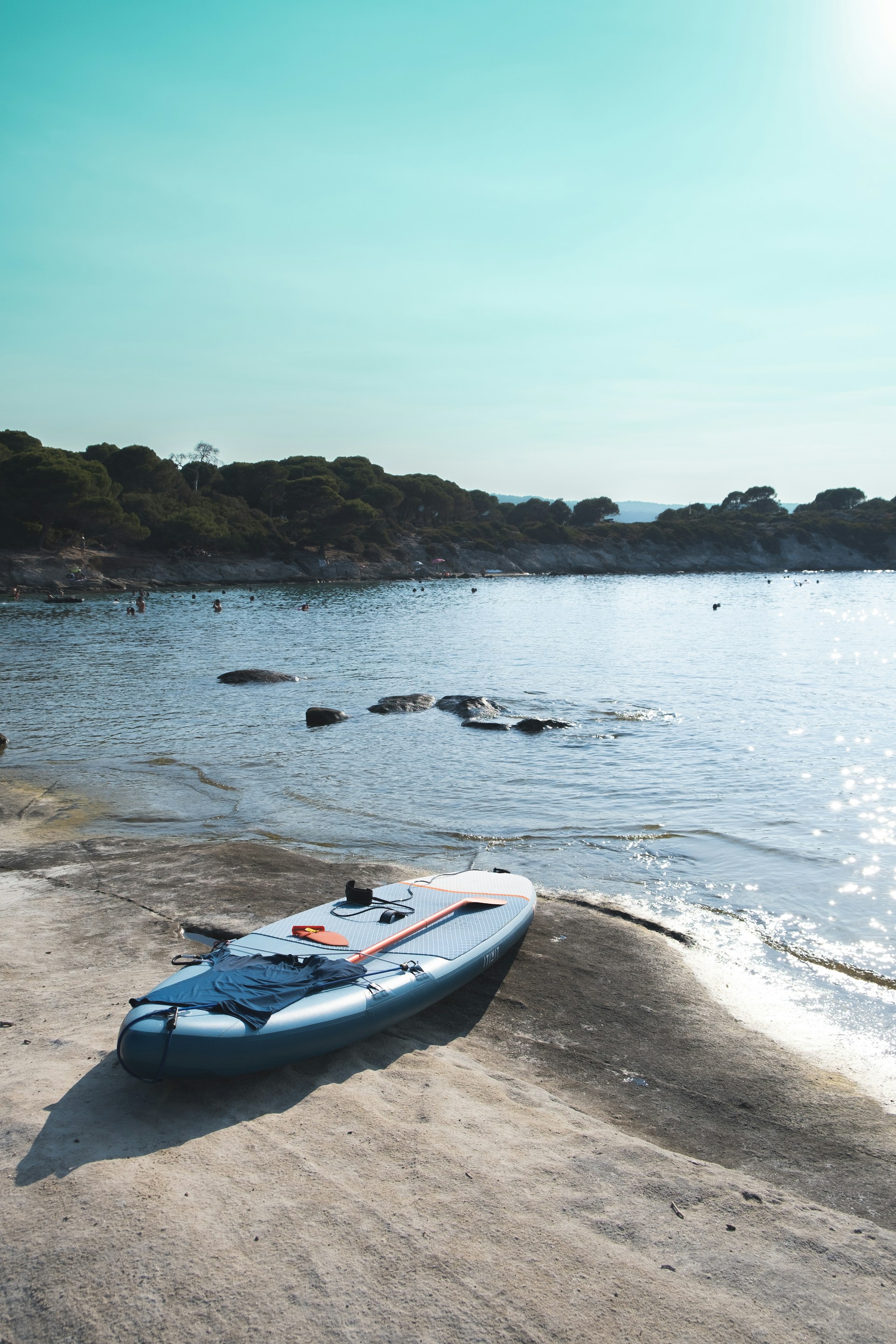 A paddleboard rests on a sandy and rocky shore next to a calm body of water. In the background, a distant shoreline with scattered greenery is visible under a clear blue sky. The sunlight glistens on the water's surface, creating a tranquil scene.
