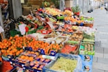 A vibrant street market stall filled with a wide variety of fresh fruits displayed in colorful boxes and crates. The produce includes oranges, apples, pears, grapes, kiwis, and other assorted fruits. Signs with prices and product information are visible.