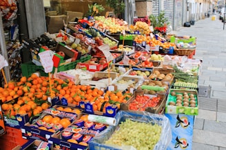 A vibrant street market stall filled with a wide variety of fresh fruits displayed in colorful boxes and crates. The produce includes oranges, apples, pears, grapes, kiwis, and other assorted fruits. Signs with prices and product information are visible.